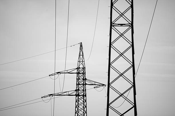 A tall electricity pylon stands silhouetted against a cloudy, overcast sky, with power lines stretching outward in multiple directions. The stark black structure contrasts with the soft gray background, highlighting the industrial design and functionality.