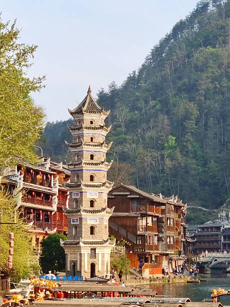 Zhangjiajie, China - Apr 07, 2025: Prominent traditional Chinese architecture of multi-tiered pagoda tower in Fenghuang Ancient Town surrounded by traditional stilt houses, colorful decorated boats and waterfront setting