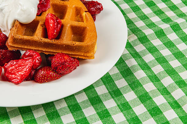 A warm waffle sits on a white plate, adorned with fresh strawberries and whipped cream, resting on a green plaid tablecloth, perfect for breakfast or brunch.