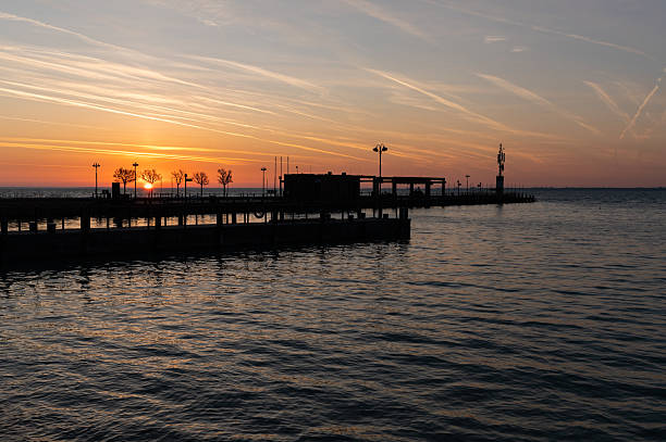 A stunning sunrise scene with a pier lined by silhouetted trees and lamp posts, reaching into the calm lake. The sky is illuminated with vibrant shades of orange and yellow, creating a peaceful and inspiring start to the day.