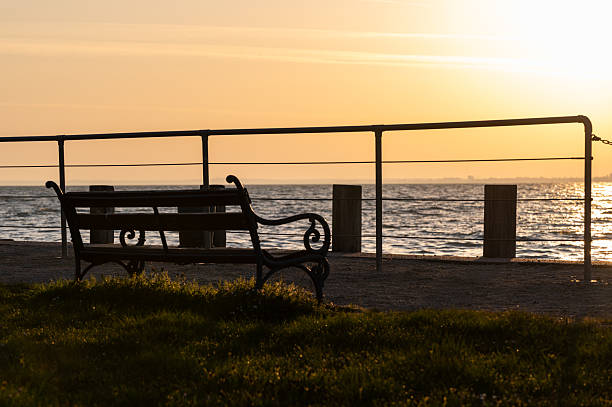 A quiet waterfront scene showing an empty bench overlooking the lake at sunrise. The golden light reflects beautifully on the water, creating a serene and contemplative atmosphere ideal for themes of relaxation and solitude.