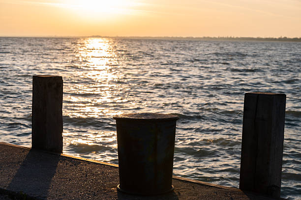 A rustic mooring post stands on a pier against the backdrop of a golden sunrise over the lake. The sunlight reflects on the rippling water, creating a warm and peaceful coastal atmosphere.
