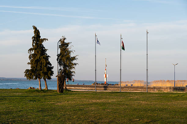 A peaceful lakeside park with neatly trimmed grass, tall trees, and several flagpoles waving in the breeze. In the background, a red and white navigation beacon stands at the water's edge under a sunny skies.
