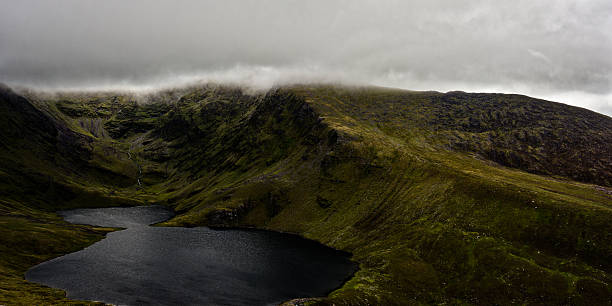 istock Coomloughra lough showcases dark waters surrounded by lush hills and dramatic clouds overhead. The tranquil setting invites exploration and admiration of nature's wonders. 2212183731