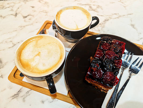 An overhead shot shows two cups of cappuccino with frothy tops and a slice of cherry cake on a black plate with two forks. The food and drinks rest on a wooden tray atop a white marble table.