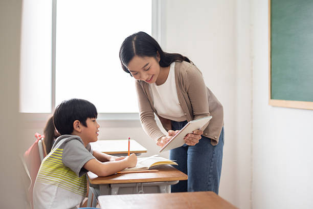 istock Smiling Asian Teacher woman assists a young boy with his studies using tablet to explain at a classroom desk while enjoying educational and learning moment. 2212192068