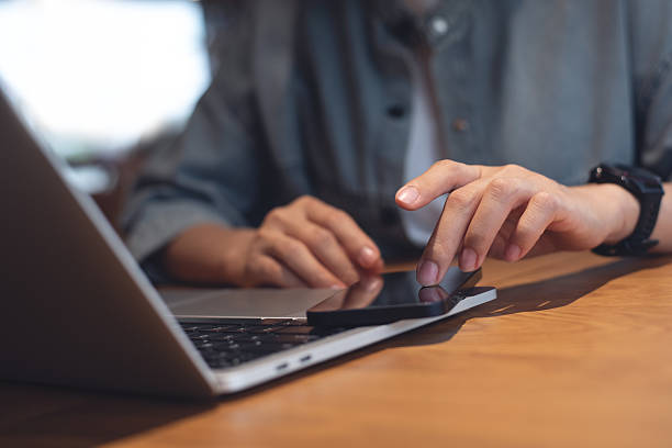 istock Woman using mobile phone surfing the internet and laptop computer on wooden table, close up. Female finger touching on smartphone screen with laptop computer on office desk, internet technology 2212193621