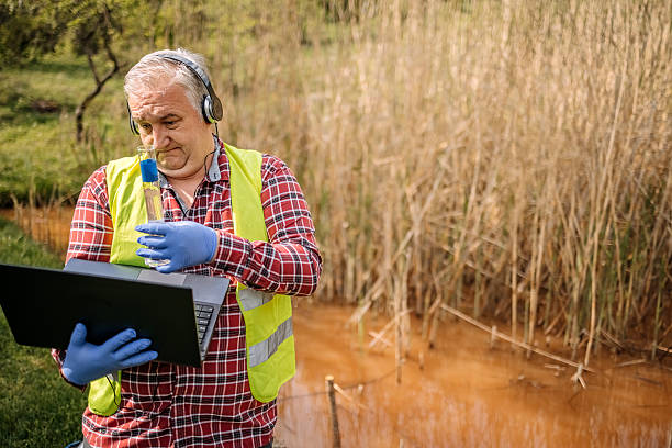 Man checking environment on mountain top, taking water samples in nature to check for pollution and communicating with colleague online using laptop