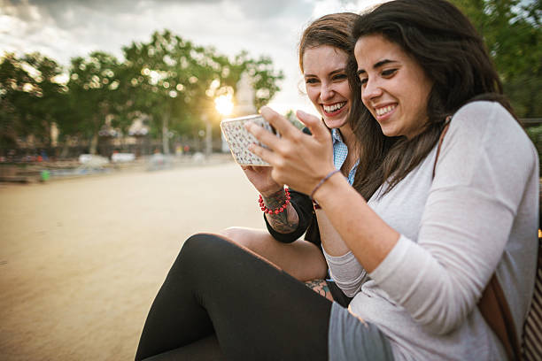 Two young women sitting outdoors, laughing together while looking at a smartphone.