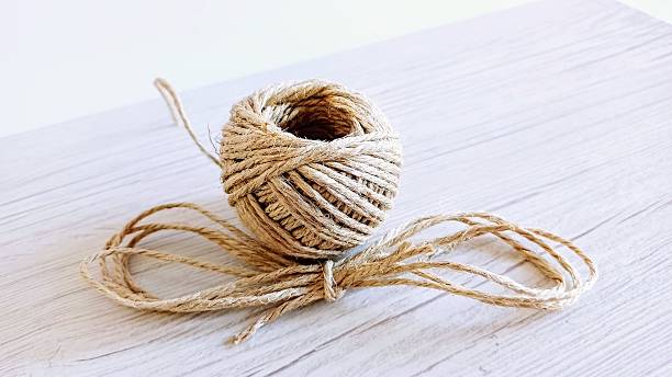A close-up of a ball of natural beige-colored twine resting on a wooden surface. The coarse texture of the twine and the subtle grain of the wood create a rustic and handmade aesthetic. A loose strand extends outward, adding a sense of natural imperfection. Perfect for concepts related to crafting, DIY projects, packaging, or eco-friendly materials.
