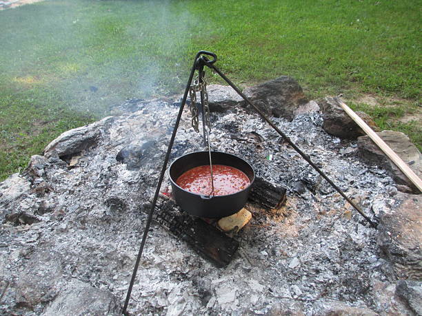 Chili cooking in Dutch oven hanging on a tripod over a smoking campfire with green grass in the background