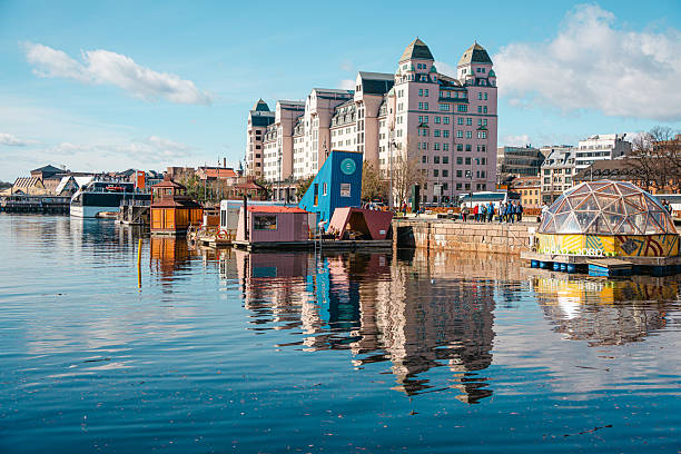 Oslo, Norway - Apr 13, 2024: Colorful floating houses and modern buildings reflect in a calm harbor under a clear blue sky.