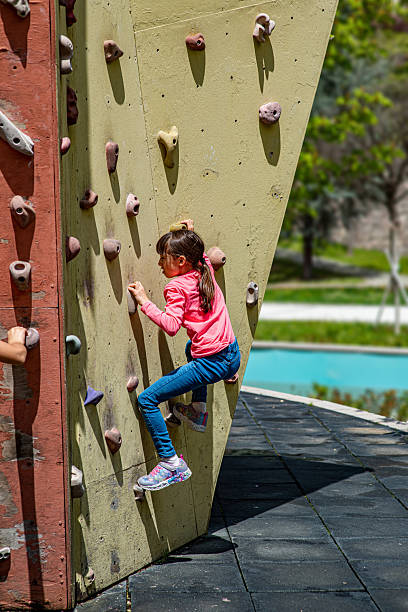 A modern indoor climbing gym featuring bouldering walls and colorful holds. A young female athlete is captured mid-action, climbing with visible focus, strength, and determination. The scene highlights themes of physical fitness, mental resilience, and an active urban lifestyle in a clean and motivating environment.
