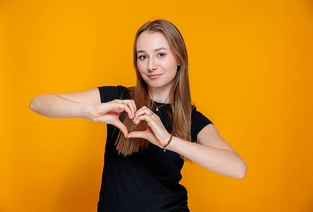 istock Smiling Young Woman Making a Heart Gesture on Orange Background 2212264645