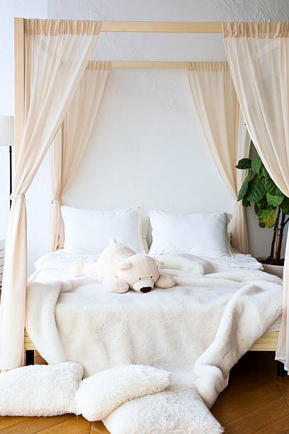 A serene bedroom features a four-poster bed draped with light fabric surrounded by cozy textiles and a plush teddy bear.