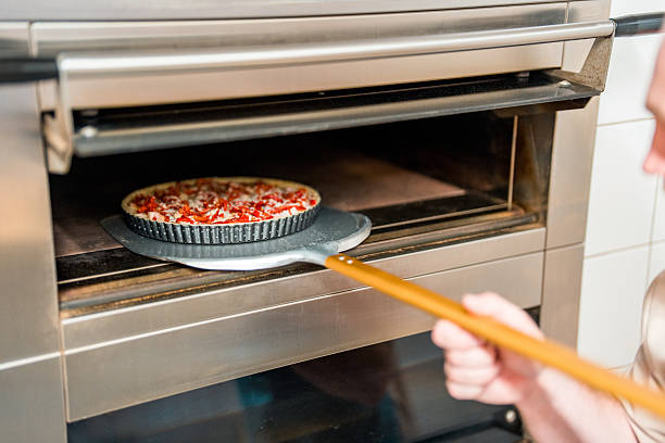 A young Caucasian baker wearing a white apron slides a freshly made pizza on a wooden peel into a stainless steel industrial oven in a professional kitchen.