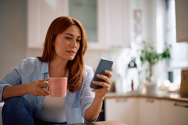 istock Mid adult woman drinking coffee while using cell phone in the kitchen. 2212274428