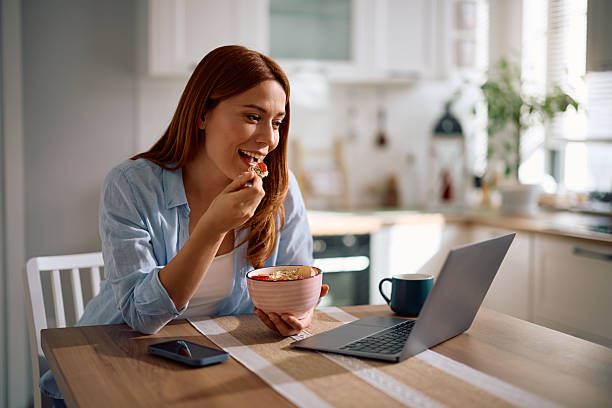 istock Happy woman eating breakfast while surfing the net on laptop at dining table. 2212274615