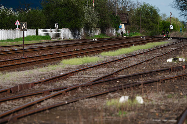 Multiple curved railway tracks stretch into the distance at a quiet rural train station, flanked by green vegetation and a white wooden fence. The scene conveys a sense of stillness and anticipation, with the rusty rails and overgrown grass hinting at the passage of time.