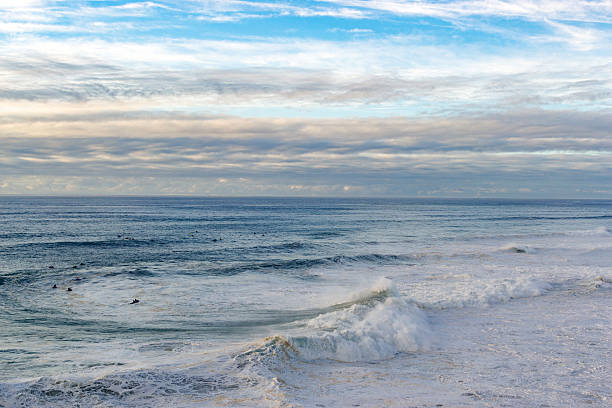 Dramatic ocean view featuring surfers, waves, and a dynamic sky. Captures the power and serenity of the sea. Perfect for travel, nature, or active lifestyle themes.