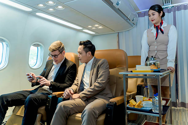 istock Businessmen discussing project on smartphone during in-flight meal service, flight attendant serving refreshments, premium travel, corporate communication scene 2212325577