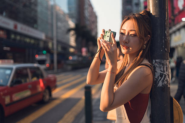 Beautiful woman with a retro camera standing outdoors at city bus station clicking moments between journey while vehicles are by the road, capturing transit life concept.