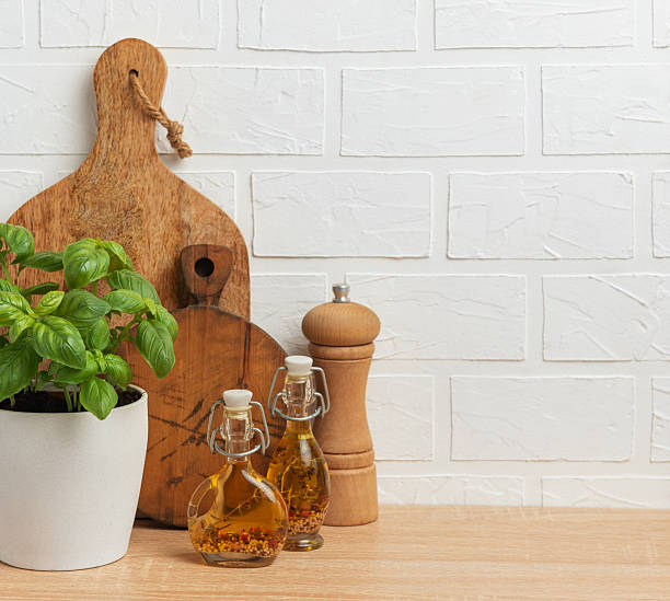 Kitchen counter with fresh basil plant, olive oil bottles, pepper mill and cutting boards against a white brick wall