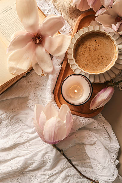 istock Lit candle in a glass jar with soft magnolia flowers, a gift box, and a coffee cup in warm pastel light on white lace fabric 2212331237