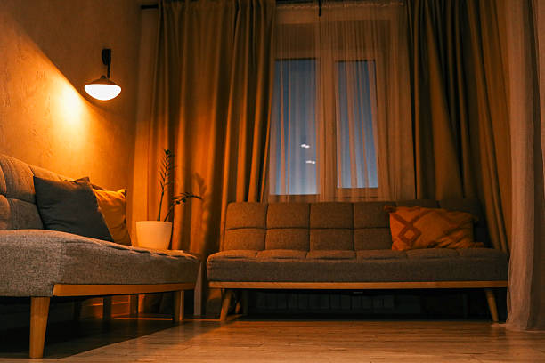 istock Cozy living room interior with warm lighting, grey sofas, decorative pillows, and a potted plant near the window during sunset 2212331890