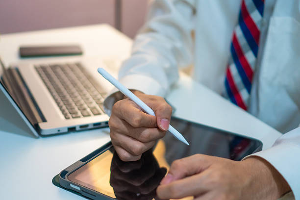 A person in formal attire using stylus on tablet, with laptop in background, suggesting professional and modern work environment