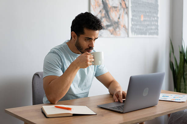 istock Focused young man drinking coffee while working on laptop at home 2212341081