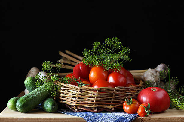 Woven basket overflowing with ripe red tomatoes takes center stage, accompanied by fresh cucumbers, garlic, and dill, creating a colorful still life against a dark backdrop