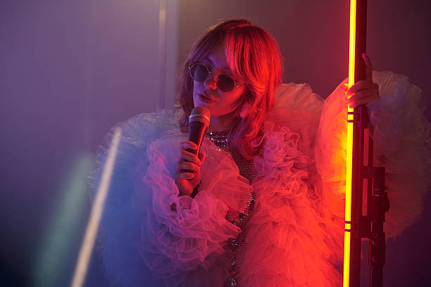 Waist up portrait of young woman holding microphone and leaning on light stand while performing during video shoot