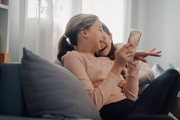 istock Two cheerful sisters laughing while watching videos on a smartphone sitting on a sofa 2212365988