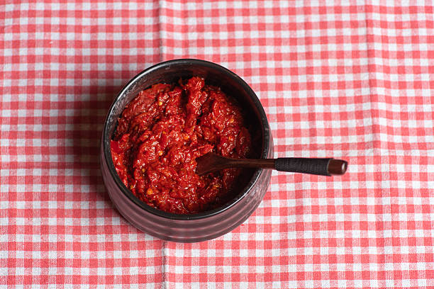 A dark bowl holds a spicy, red pepper paste with a wooden spoon, resting on a checker pattern table surface, emphasizing the rich, fiery color and texture of the flavorful condiment.