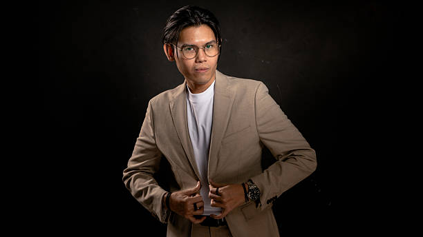 istock Asian man in beige blazer and white t-shirt adjusting jacket while looking sideways in studio with dark background, expressing confidence and style, ideal for business, fashion, or branding use 2212386948