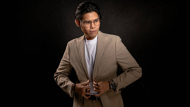 istock Asian man in beige blazer and white t-shirt adjusting jacket while looking sideways in studio with dark background, expressing confidence and style, ideal for business, fashion, or branding use 2212387028