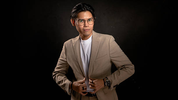istock Asian man in beige blazer and white t-shirt adjusting jacket while looking sideways in studio with dark background, expressing confidence and style, ideal for business, fashion, or branding use 2212387030
