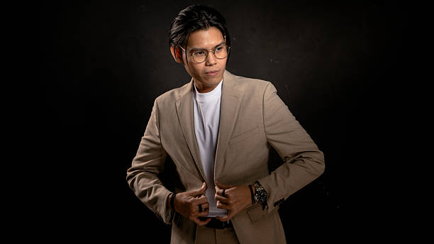 istock Asian man in beige blazer and white t-shirt adjusting jacket while looking sideways in studio with dark background, expressing confidence and style, ideal for business, fashion, or branding use 2212387031