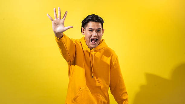 istock Asian man in bright yellow hoodie raising both hands with open palms and playful expression, smiling widely against vibrant yellow background. Energetic gesture showing excitement or fun 2212389130