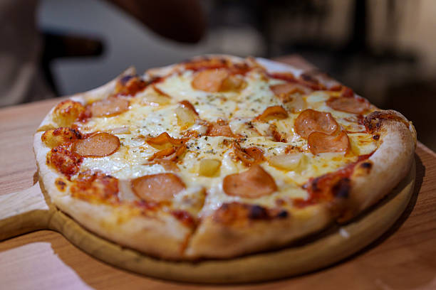 A freshly baked cheese pizza with pepperoni slices and pineapple served on a wooden tray in a restaurant.