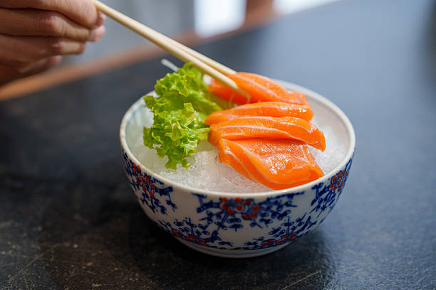 A woman using chopsticks to pick a slice of fresh salmon sashimi from a beautifully arranged bowl.