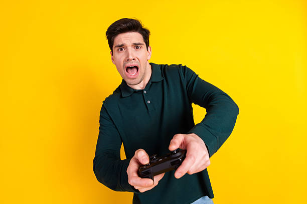 istock Young man in green polo shirt holding game controller with surprised expression on yellow background 2212403981