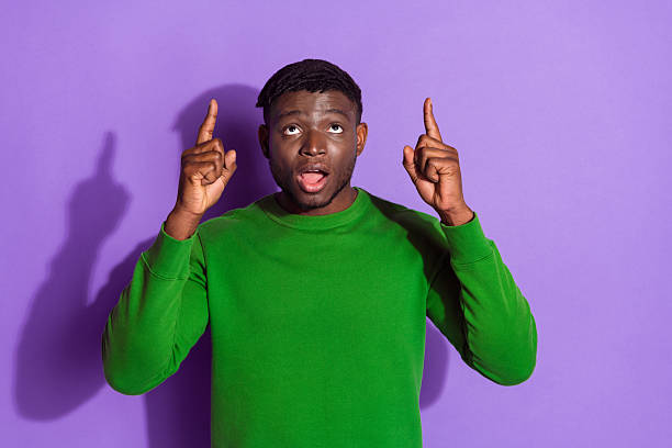 istock Young man in a green sweatshirt pointing upwards with an expressive gesture against a vibrant purple background 2212406921