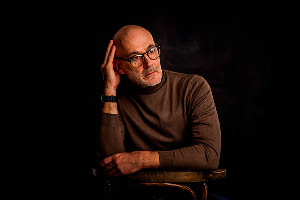 A senior man sitting pensively against a dark background. Confident male wearing reading glasses and sweater. Isolated background. Copy space.