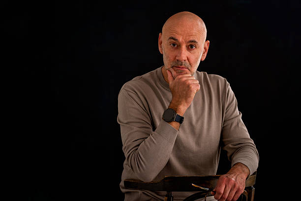 A senior man sitting pensively against a dark background. Confident male wearing sweater. Isolated background. Copy space.