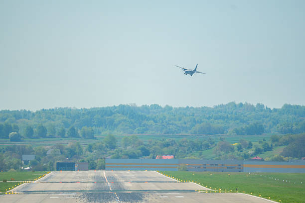 A military aircraft lands above the runway, framed by lush greenery and bright blue skies in daylight.