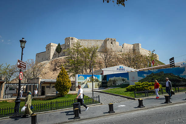 Istanbul, Turkey - April 18, 2025: The historic Gaziantep Castle, a significant landmark in Gaziantep, Turkey. The ancient stone fortress stands atop a hill, overlooking the city, symbolizing centuries of history. In the foreground, a modern monument and urban scenery contrast with the castle’s timeless architecture. People can be seen walking nearby, and vehicles line the streets, adding to the vibrant atmosphere. The clear blue sky and scattered clouds provide a dynamic backdrop, enhancing the overall aesthetic of the city. This photograph highlights the fusion of heritage and modern life in Gaziantep.