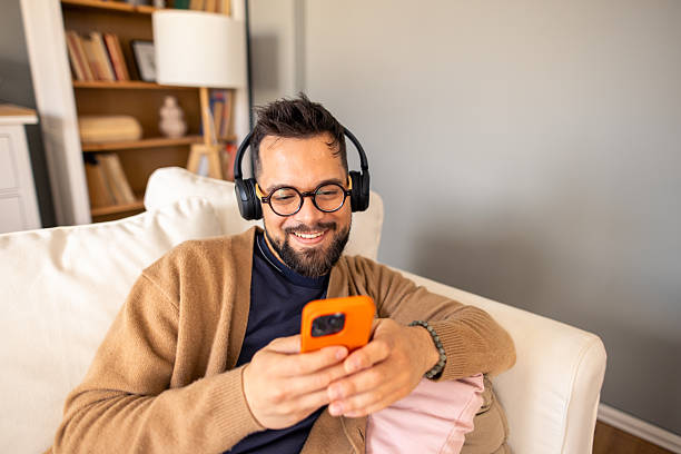 istock Caucasian Man Smiling While Relaxing on Couch Wearing Headphones and Using Smartphone 2212432796