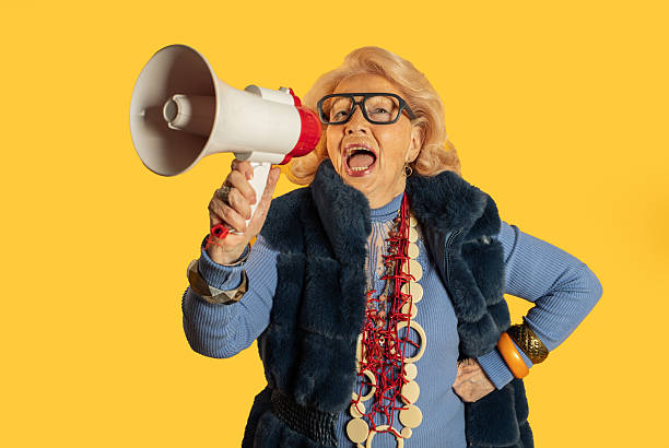 istock Senior woman shouting through megaphone on yellow background 2212433850